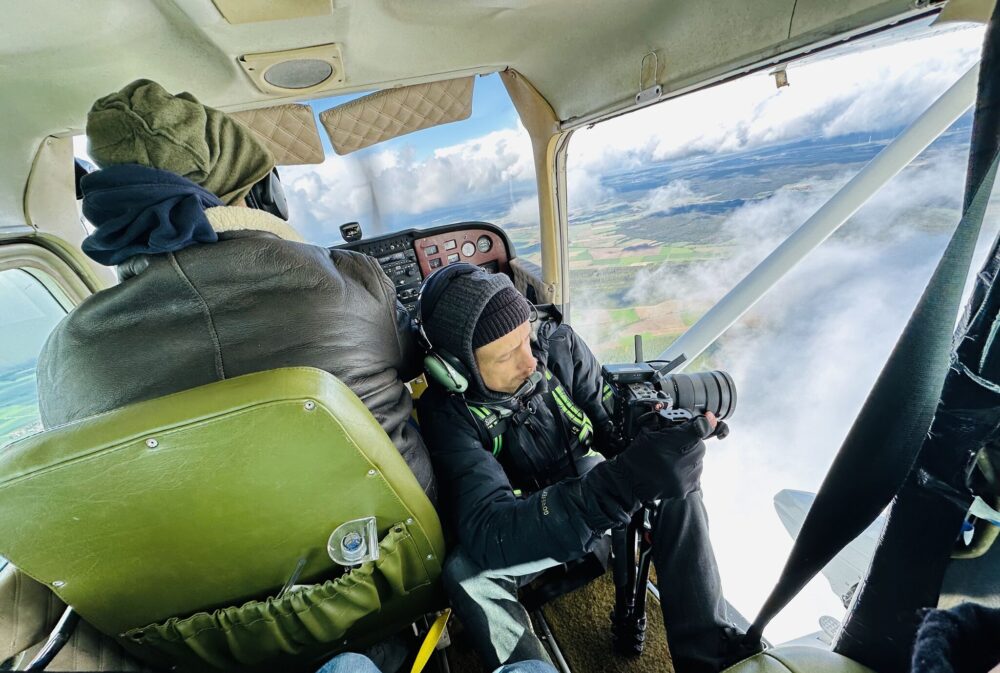 Ein Kameramann sitzt an der offenen Tür eines Kleinflugzeuges über den Wolken.