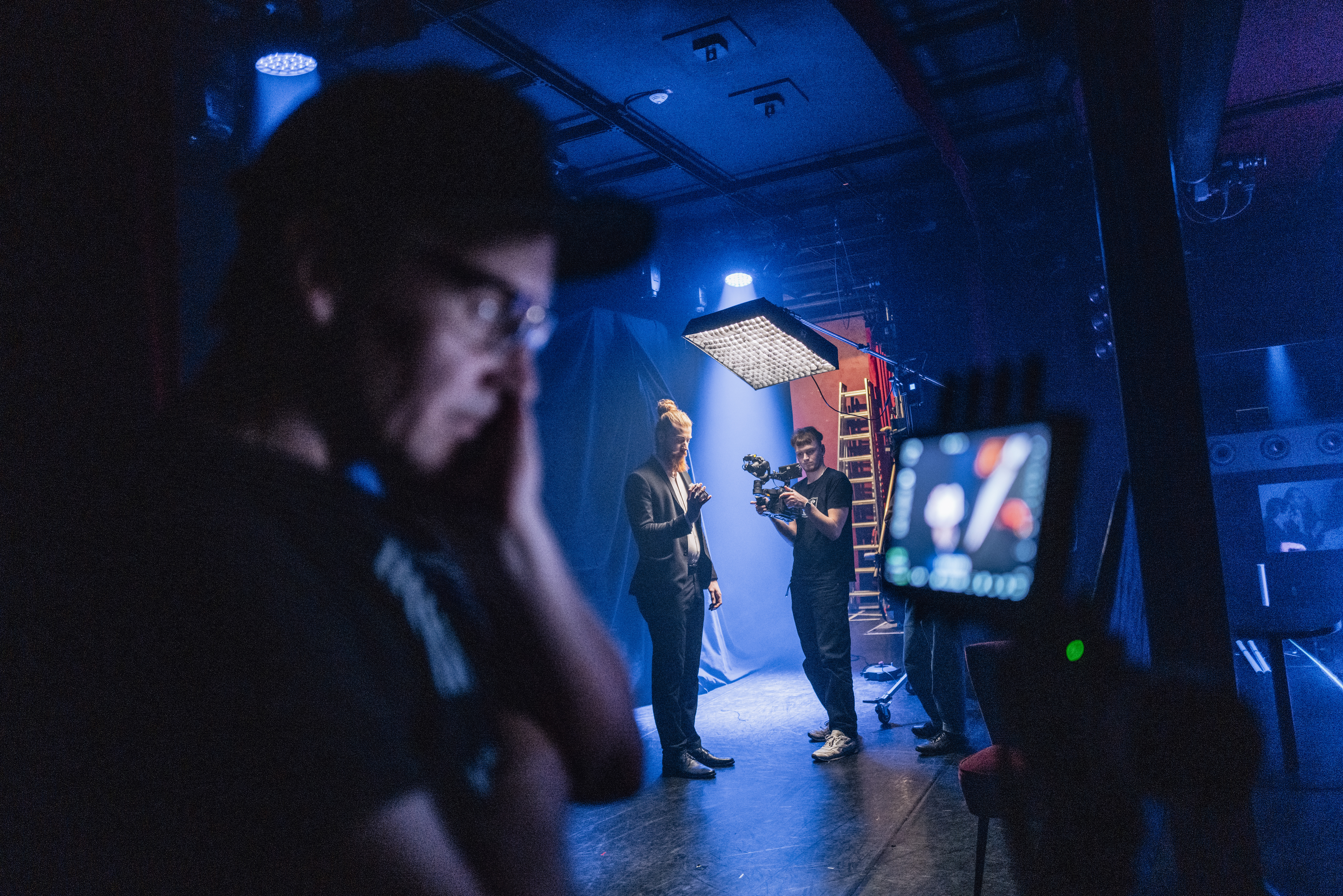 Ein Regisseur betrachtet einen Monitor in einem dunklen Filmset. Im Hintergrund filmt ein Kameramann einen Zauberer mit Spielkarten in der Hand.