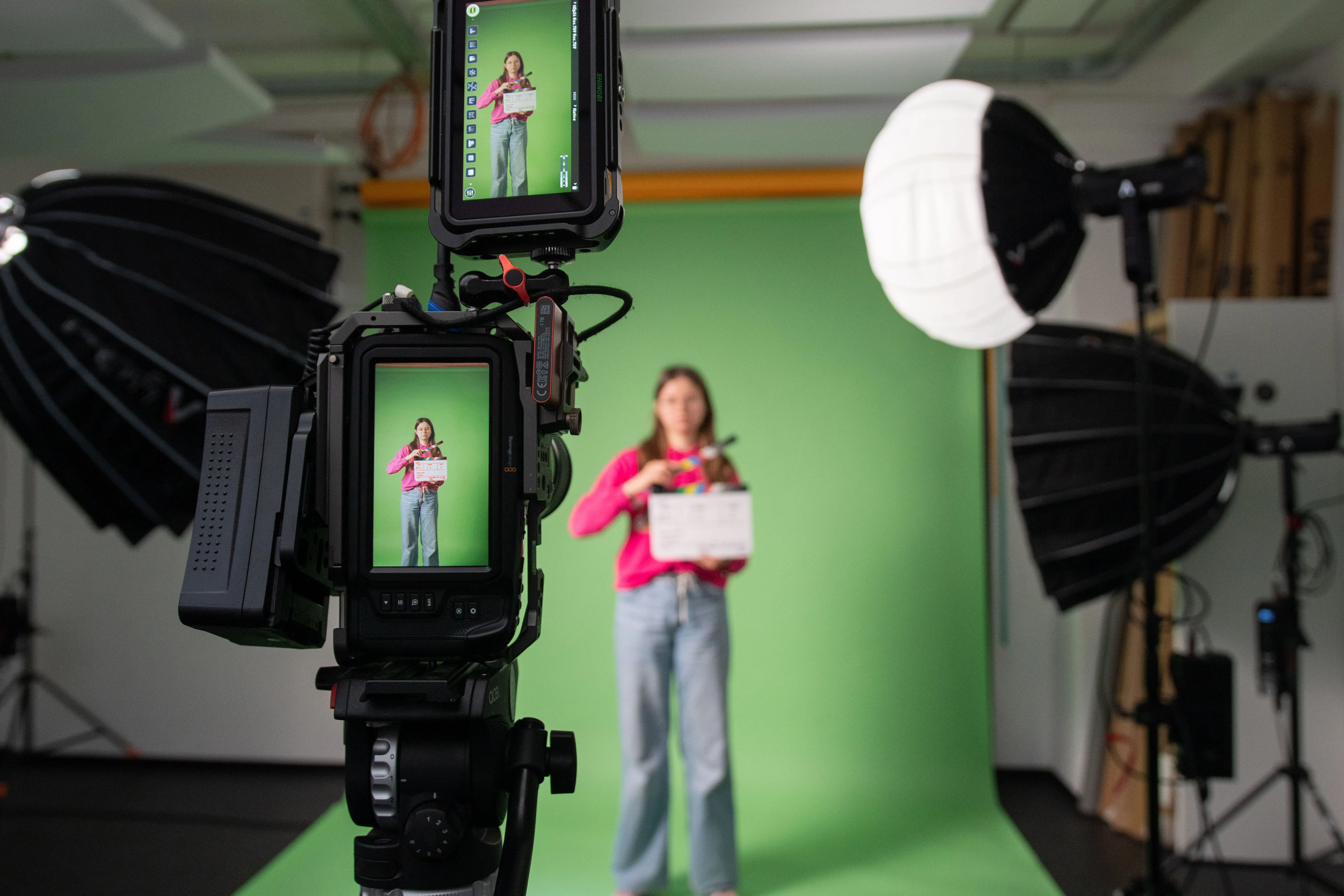 Eine Person in der Unschärfe vor einem Greenscreen, im Vordergrund scharf eine Kamera.