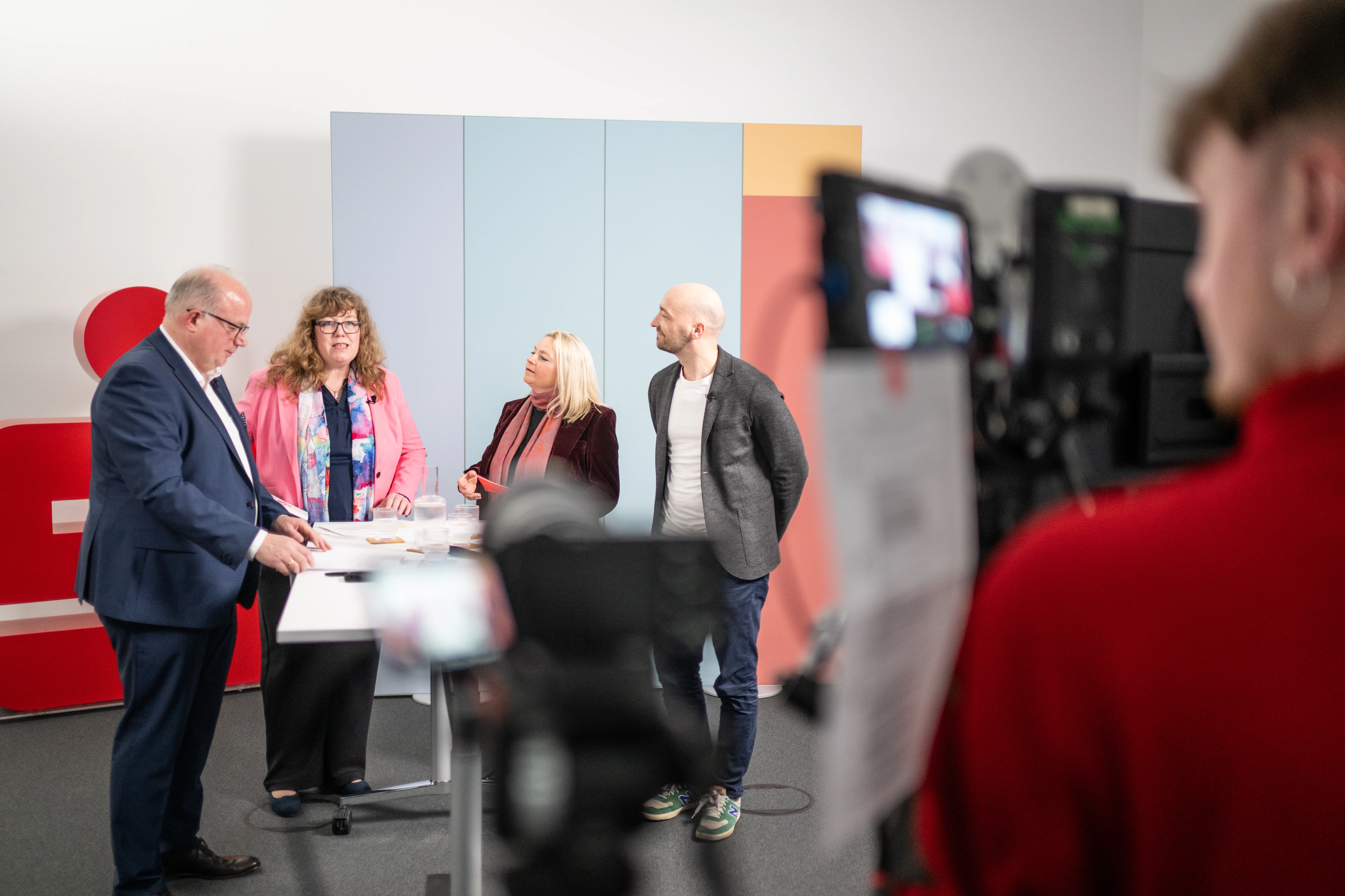 Eine Gesprächsrunde für die Mitarbeiter der Sparkasse Hannover bei einer Livestream-Produktion. Im Vordergrund rechts ist unscharf ein Kameramann an einer Kamera zu erkennen. Im linken Bildteil stehen 4 Personen an einem Tisch und unterhalten sich.