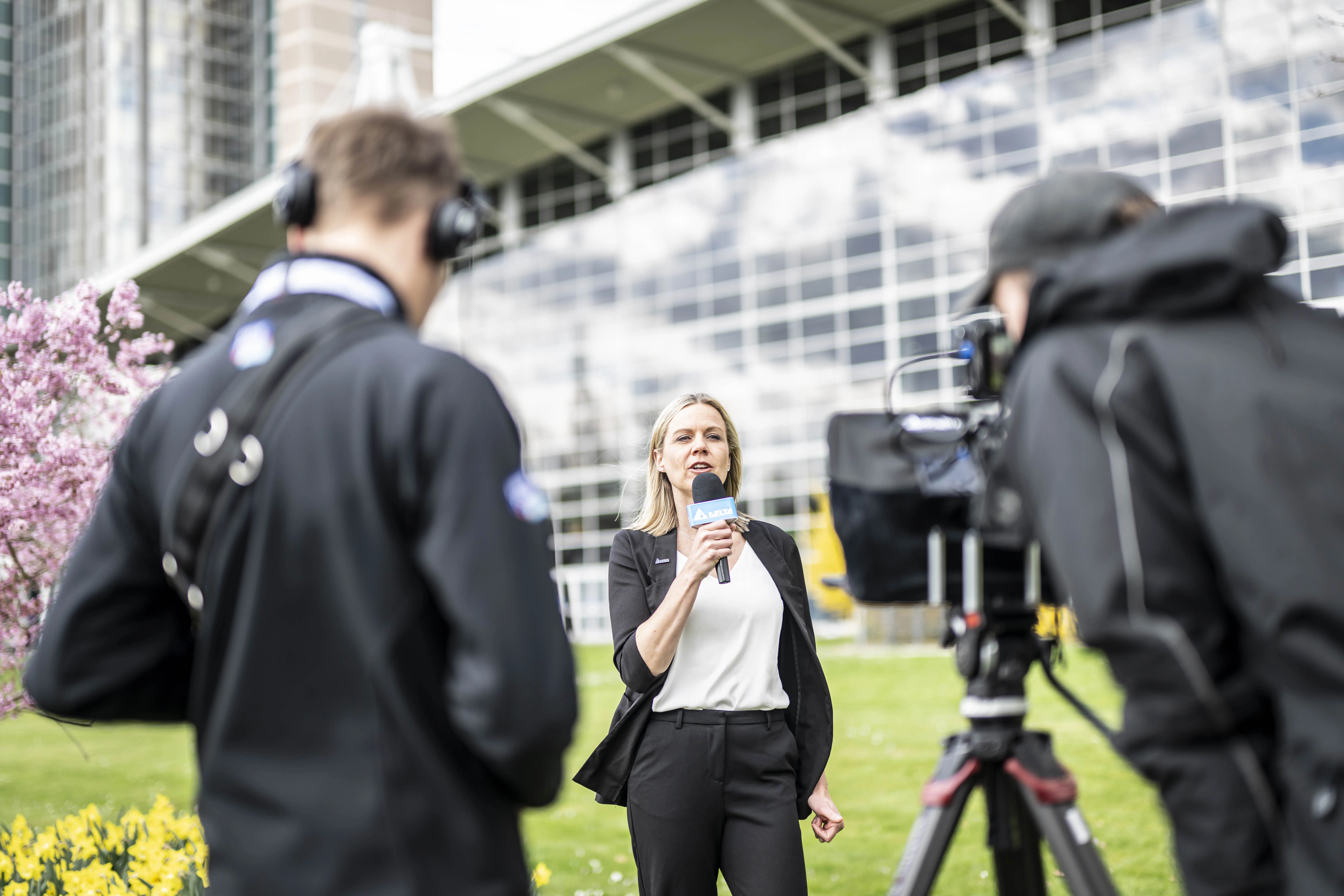 Eine Moderatorin mit einem Mikrofon vor einer Messehalle in Hannover. Im Vordergrund zwei Kameraleute.