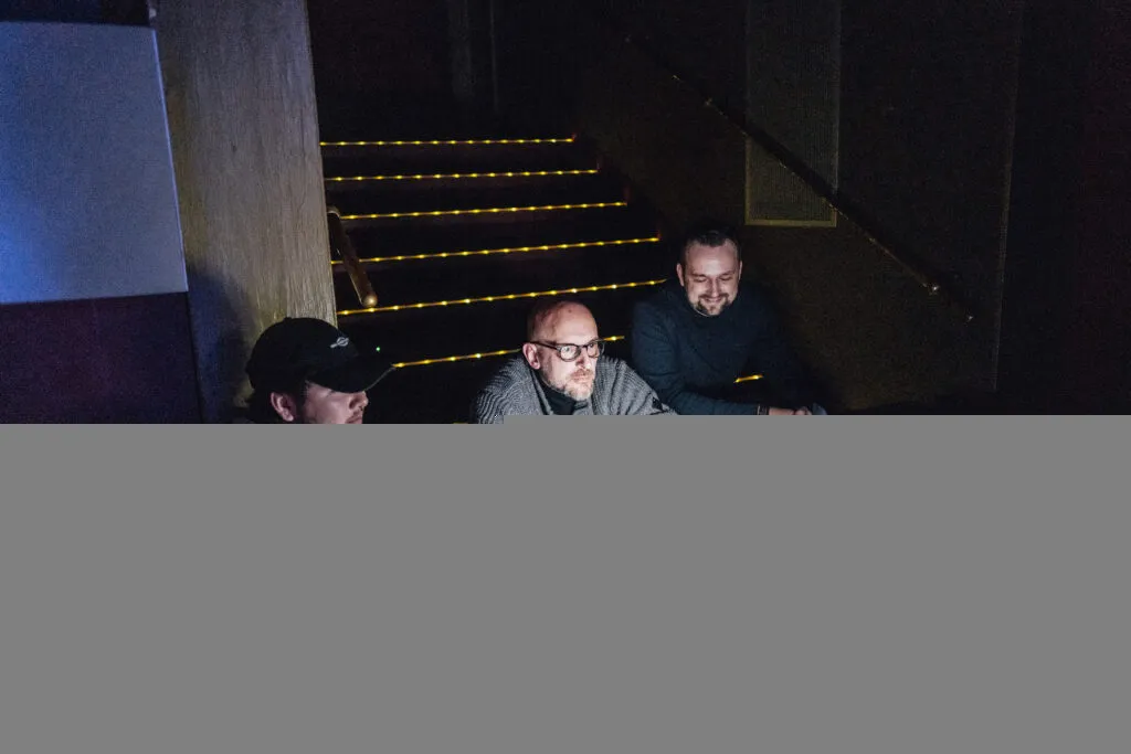 Ein Regisseur und sein Team sitzen auf einer Treppe bei einem Filmset und betrachteten einen Monitor.