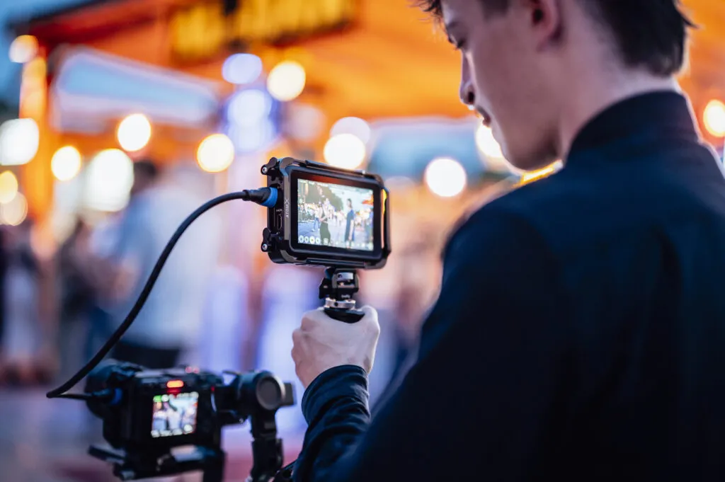 Ein Auszibildender für Mediengestalter Bild und Ton hält eine Kamera beim Filmen in der Hand. Im Hintergrund sind die unscharfen Lichter eines Events.