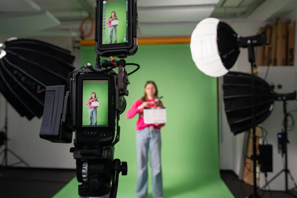 Eine Person in der Unschärfe vor einem Greenscreen, im Vordergrund scharf eine Kamera.