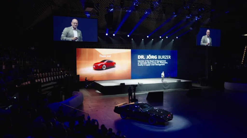 In einer Messehalle steht ein Auto für ein Event. Auf der Bühne findet eine Moderation statt.