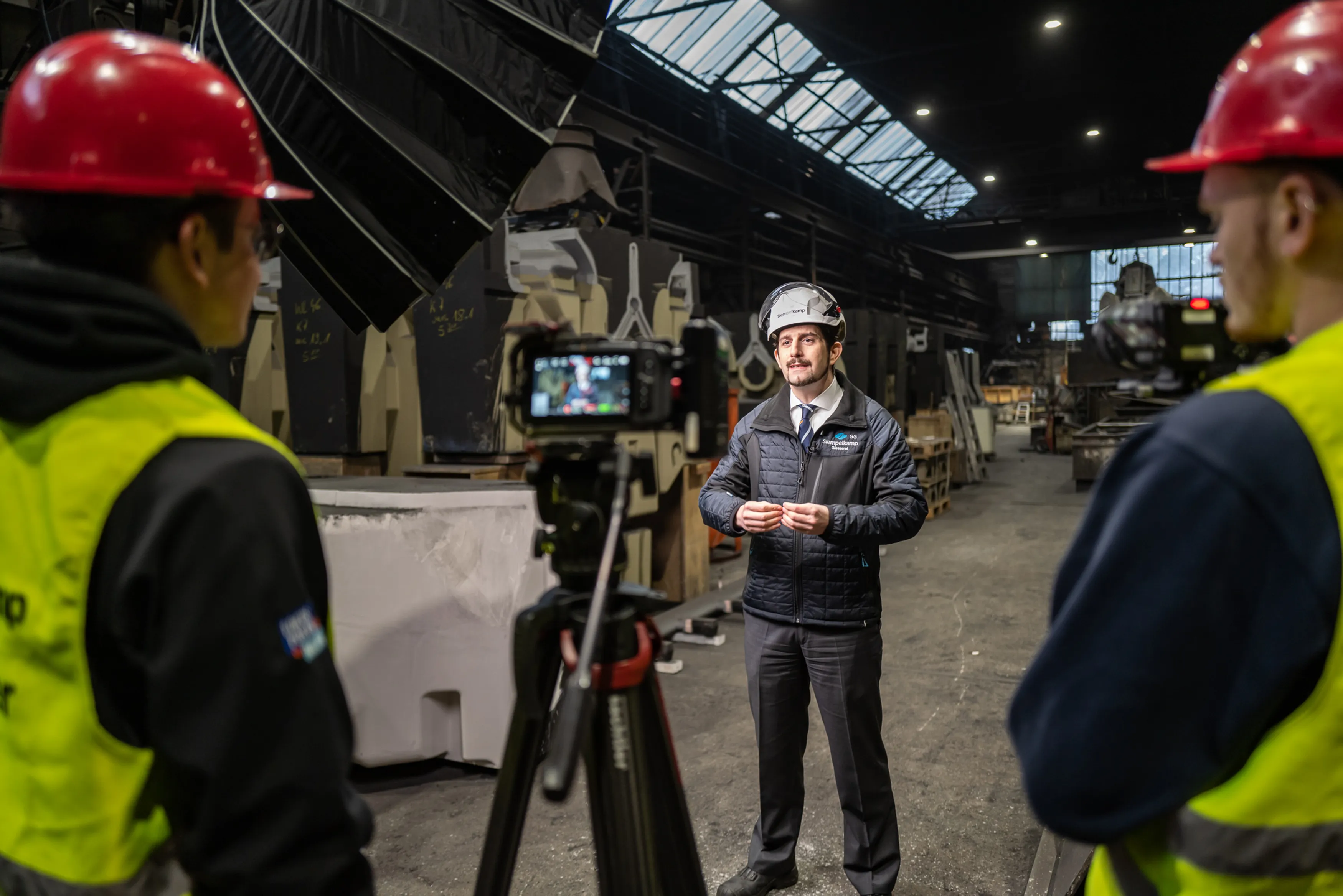 Ein Geschäftsführer einer Firma steht in der Produktionshalle und wird bei einem Interview gefilmt. Im Vordergrund stehen zwei Personen neben einer Kamera.
